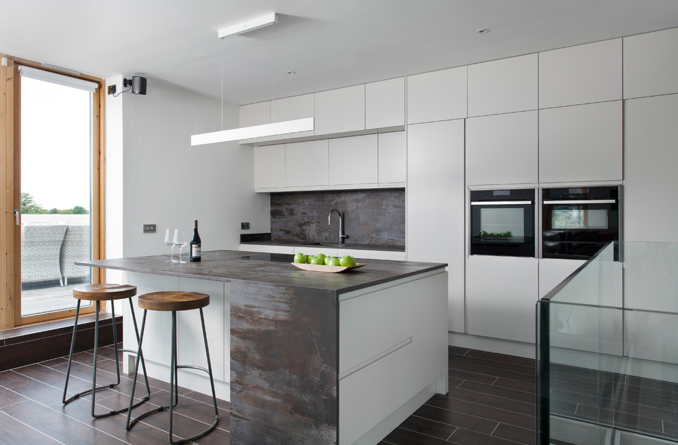 Large contemporary single-wall kitchen/diner in Dublin with a built-in sink, flat-panel cabinets, grey cabinets, composite countertops, brown splashback, stone slab splashback, black appliances, ceramic flooring, an island, brown floors and brown worktops.
