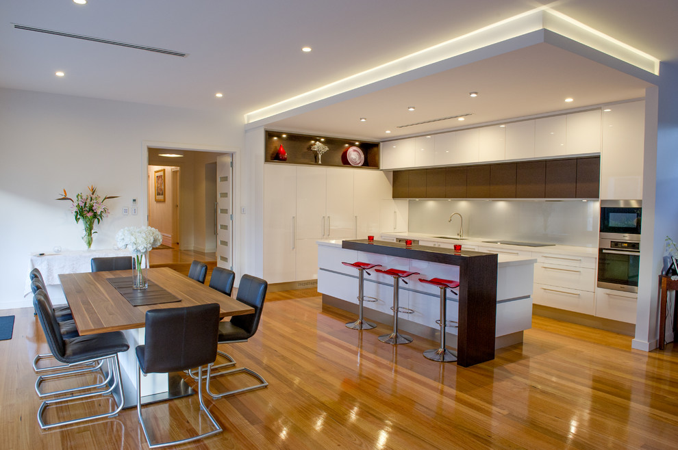 Moderne Wohnküche in L-Form mit Unterbauwaschbecken, flächenbündigen Schrankfronten, weißen Schränken, Glasrückwand, Küchengeräten aus Edelstahl, braunem Holzboden und Kücheninsel in Adelaide