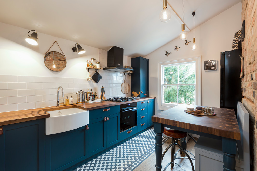 Mid-sized trendy galley ceramic tile kitchen photo in London with a farmhouse sink, shaker cabinets, blue cabinets, wood countertops, beige backsplash, subway tile backsplash, black appliances and an island