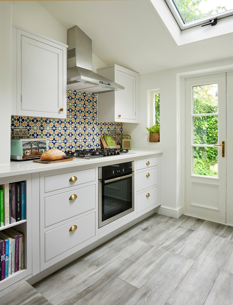 Cette photo montre une cuisine chic avec un placard à porte shaker, des portes de placard blanches, une crédence multicolore, un électroménager en acier inoxydable, un sol gris et un plan de travail blanc.