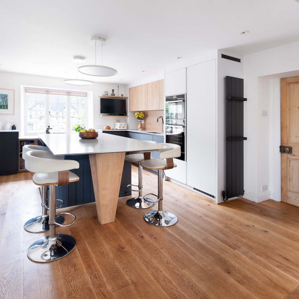 Inspiration for a medium sized modern u-shaped open plan kitchen in Gloucestershire with a built-in sink, flat-panel cabinets, composite countertops, wood splashback, black appliances, an island, beige floors and white worktops.