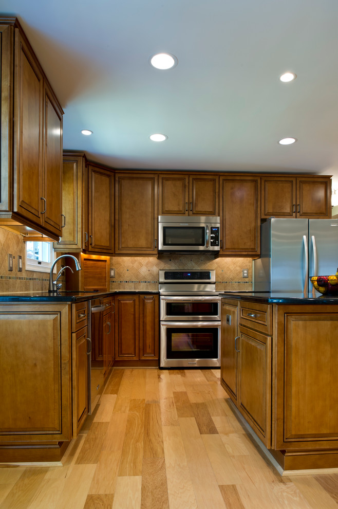 Example of a mid-sized classic l-shaped light wood floor open concept kitchen design in DC Metro with an undermount sink, raised-panel cabinets, medium tone wood cabinets, granite countertops, beige backsplash, stone tile backsplash, stainless steel appliances and an island