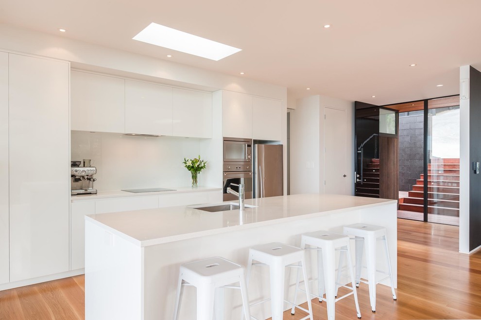 Inspiration for a large contemporary galley kitchen in Auckland with white splashback, stainless steel appliances, an island, white worktops, a submerged sink, flat-panel cabinets, white cabinets, medium hardwood flooring and brown floors.