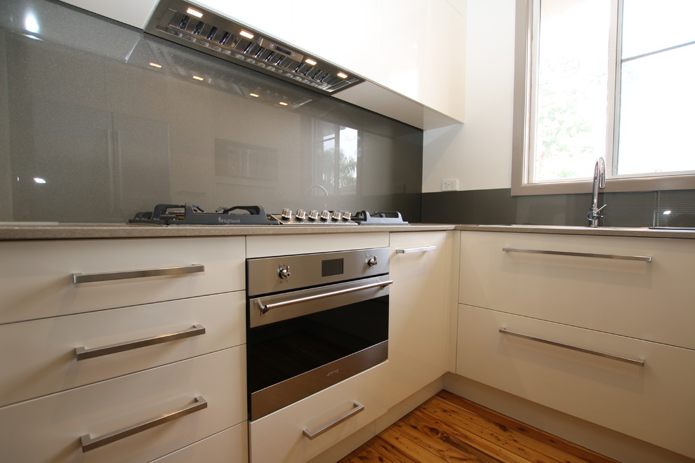 Modern inredning av ett litet kök, med en dubbel diskho, släta luckor, beige skåp, bänkskiva i kvarts, stänkskydd med metallisk yta, glaspanel som stänkskydd, rostfria vitvaror och ljust trägolv