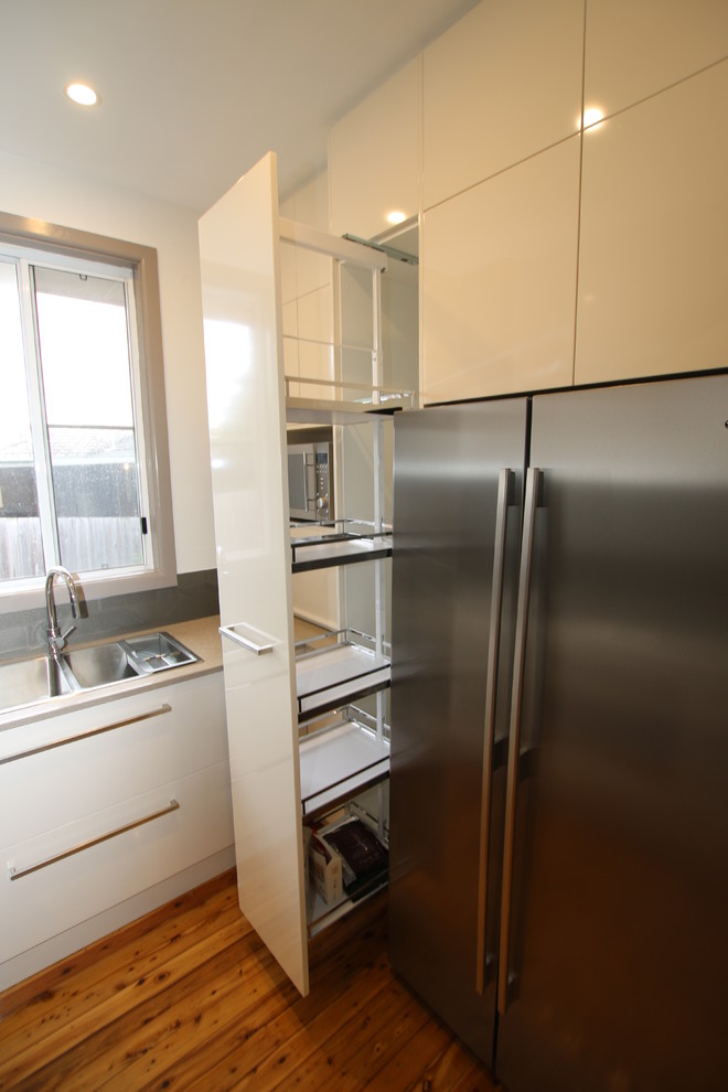 This is an example of a small contemporary u-shaped kitchen pantry in Sydney with a double-bowl sink, flat-panel cabinets, beige cabinets, engineered stone countertops, metallic splashback, glass sheet splashback, stainless steel appliances, light hardwood flooring and no island.