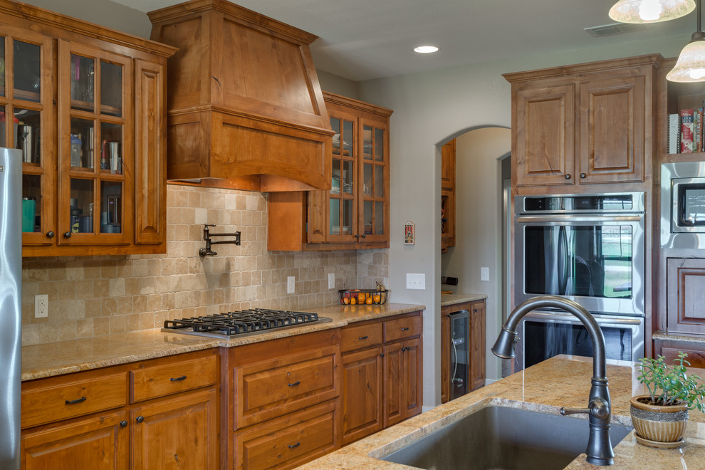 Rustik inredning av ett mellanstort kök, med en undermonterad diskho, luckor med upphöjd panel, skåp i mellenmörkt trä, granitbänkskiva, beige stänkskydd, stänkskydd i travertin, rostfria vitvaror, betonggolv, en köksö och brunt golv