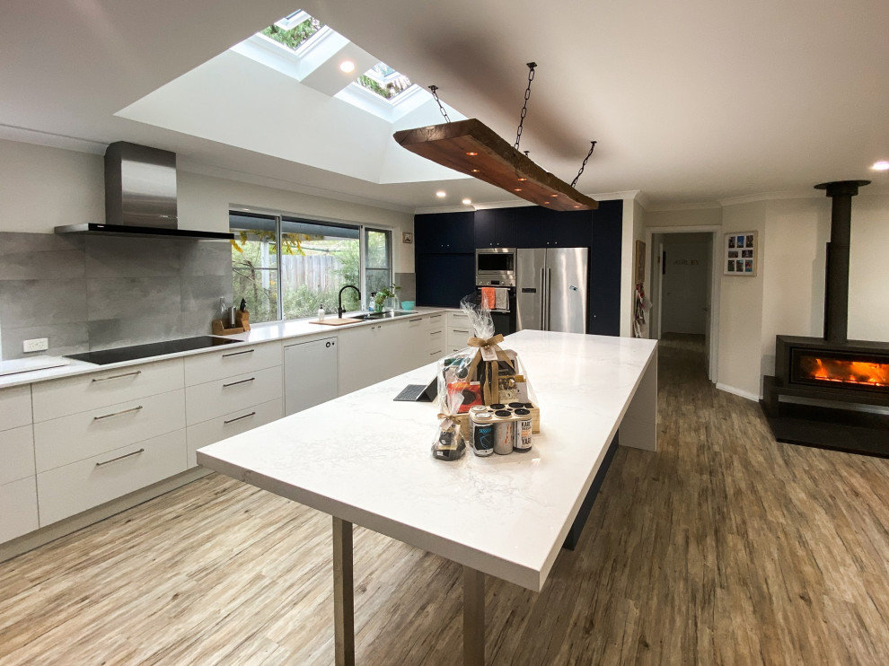 Large modern galley open plan kitchen in Other with a double-bowl sink, flat-panel cabinets, white cabinets, composite countertops, grey splashback, porcelain splashback, stainless steel appliances, laminate floors, an island, beige floors and white worktops.