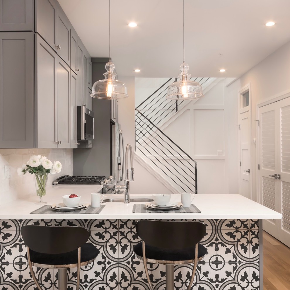 This is an example of a small traditional l-shaped open plan kitchen in DC Metro with a single-bowl sink, shaker cabinets, grey cabinets, engineered stone countertops, white splashback, ceramic splashback, stainless steel appliances, medium hardwood flooring, a breakfast bar and brown floors.