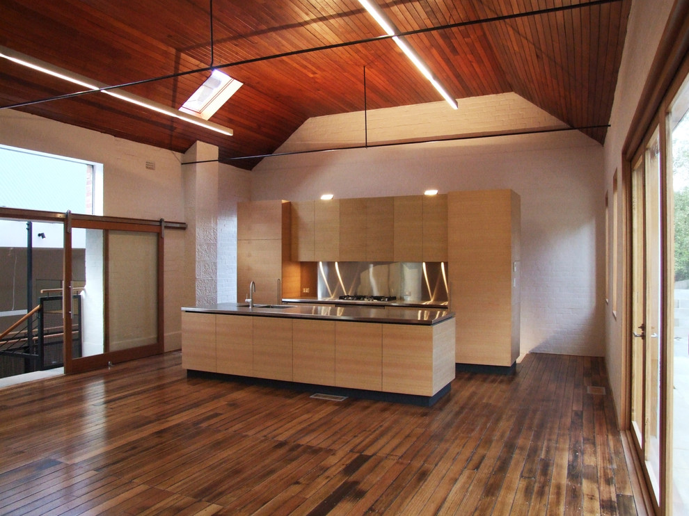Cette image montre une grande cuisine ouverte linéaire et encastrable design en bois clair avec un évier encastré, un placard à porte plane, un plan de travail en inox, une crédence grise, un sol en bois brun et îlot.