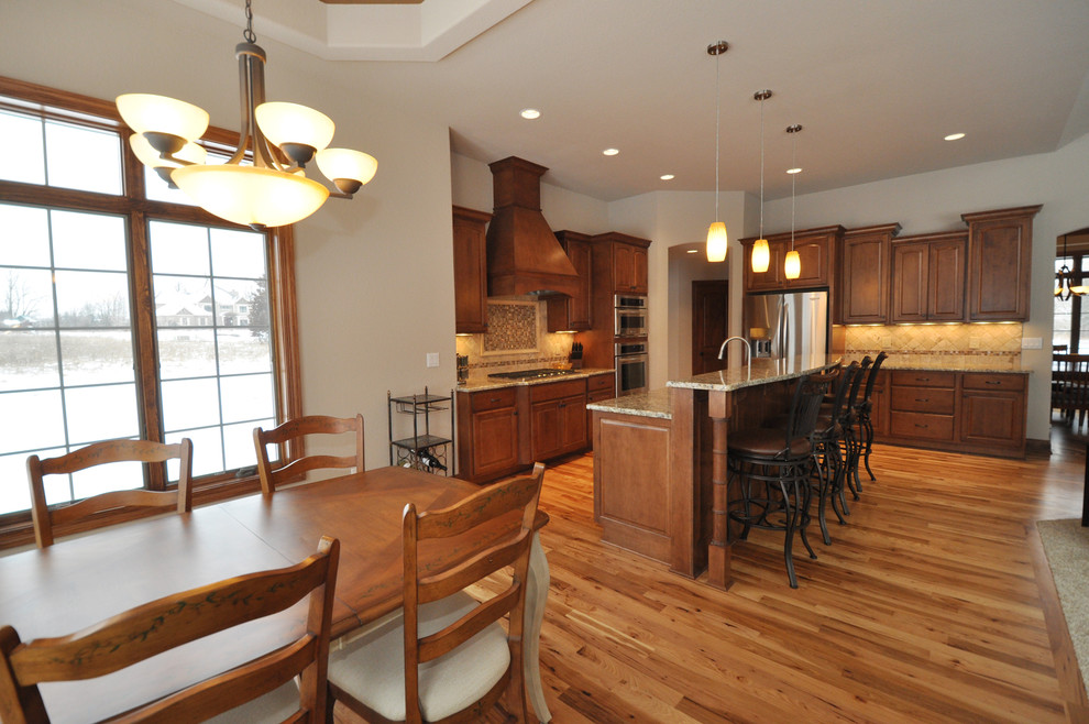 Eat-in kitchen - large traditional l-shaped medium tone wood floor eat-in kitchen idea in Milwaukee with raised-panel cabinets, medium tone wood cabinets, granite countertops, beige backsplash, ceramic backsplash, stainless steel appliances and an island
