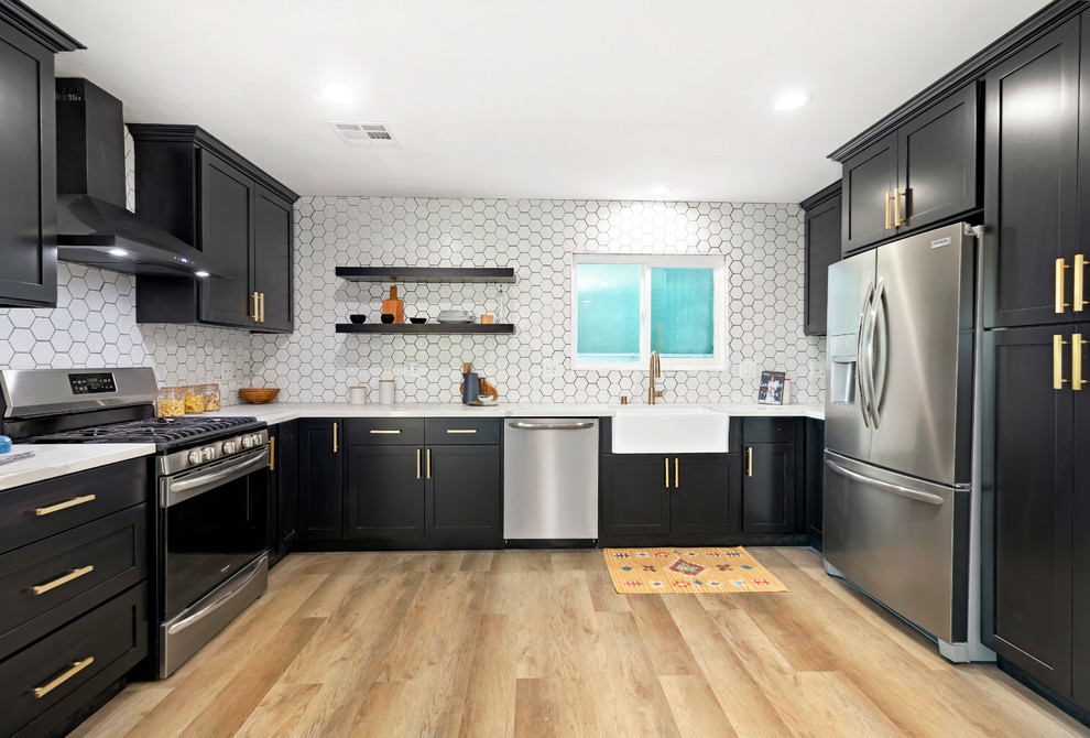 Photo of a traditional u-shaped kitchen in Los Angeles with a belfast sink, shaker cabinets, black cabinets, white splashback, stainless steel appliances, medium hardwood flooring, brown floors and white worktops.