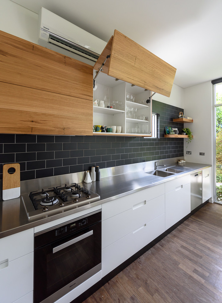 Mid-sized minimalist galley medium tone wood floor and brown floor open concept kitchen photo in Melbourne with an integrated sink, flat-panel cabinets, white cabinets, stainless steel countertops, black backsplash, ceramic backsplash, stainless steel appliances and an island
