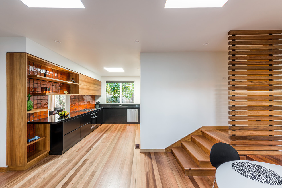 Modern inredning av ett kök och matrum, med en undermonterad diskho, släta luckor, svarta skåp, bänkskiva i kvarts, orange stänkskydd, stänkskydd i cementkakel, rostfria vitvaror och mellanmörkt trägolv