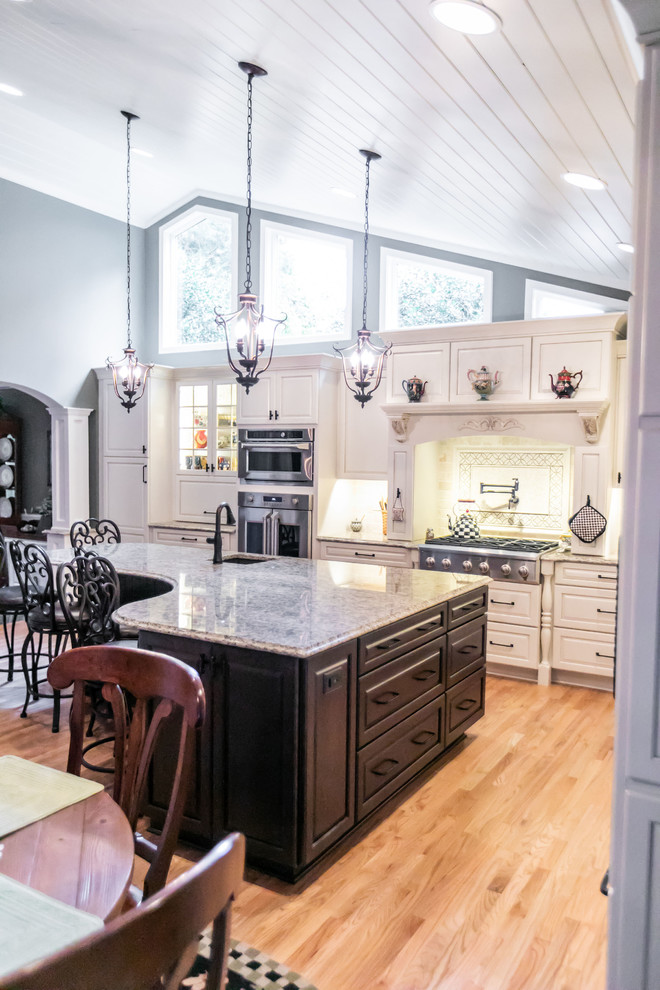 Eat-in kitchen - large traditional l-shaped light wood floor eat-in kitchen idea in Baltimore with a farmhouse sink, raised-panel cabinets, white cabinets, granite countertops, beige backsplash, travertine backsplash, paneled appliances, an island and brown countertops