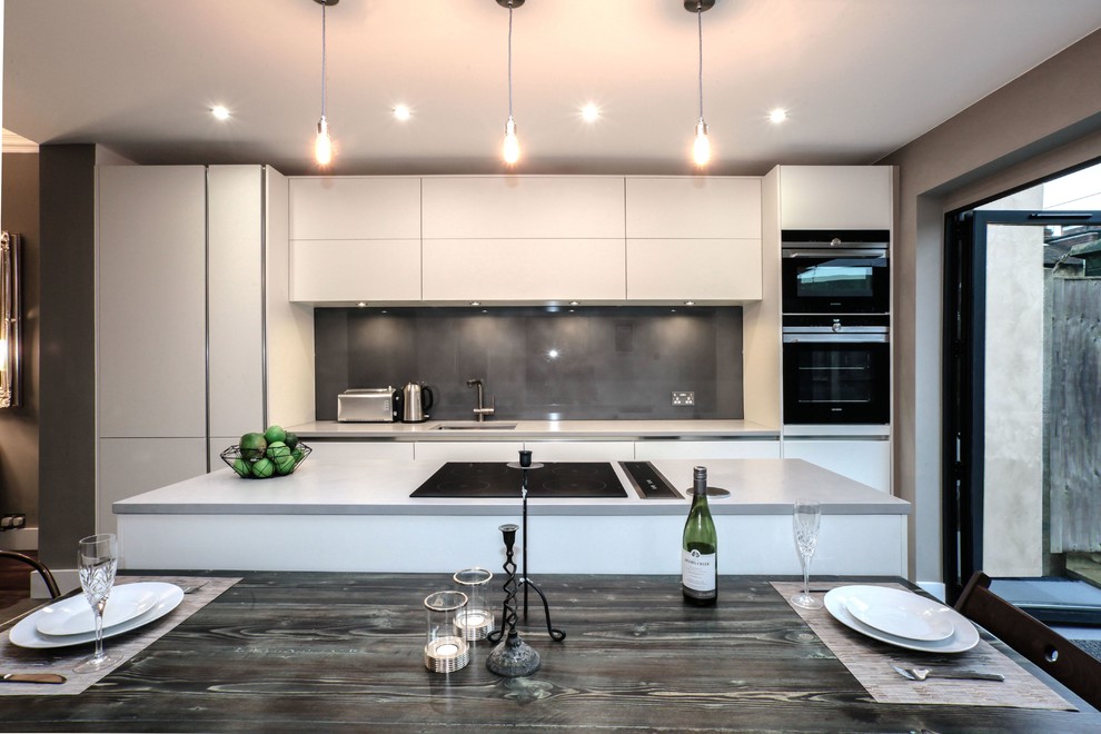 Photo of a large modern galley kitchen/diner in London with a submerged sink, flat-panel cabinets, white cabinets, composite countertops, grey splashback, glass sheet splashback, black appliances, ceramic flooring and an island.