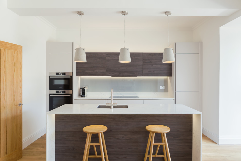 Example of a trendy light wood floor and beige floor kitchen design in London with an undermount sink, flat-panel cabinets, dark wood cabinets, gray backsplash, glass sheet backsplash, stainless steel appliances, an island and white countertops