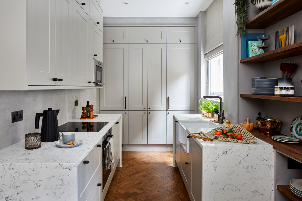 Contemporary u-shaped kitchen in London with a belfast sink, shaker cabinets, white cabinets, grey splashback, black appliances, dark hardwood flooring, brown floors and white worktops.