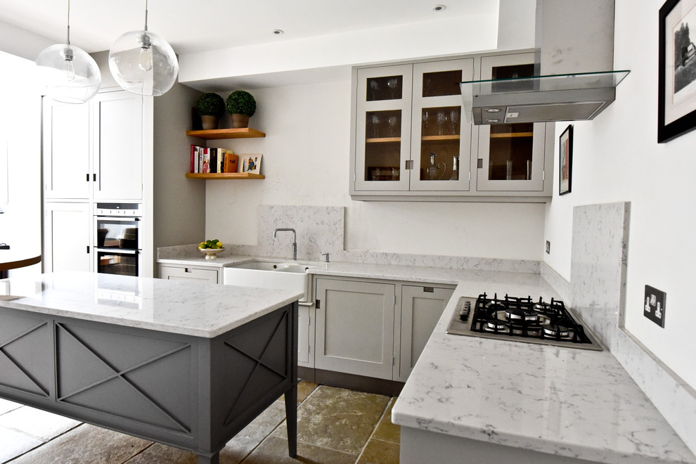 Photo of a large modern galley kitchen/diner in London with a belfast sink, grey cabinets, marble worktops, white splashback, an island, glass-front cabinets and integrated appliances.
