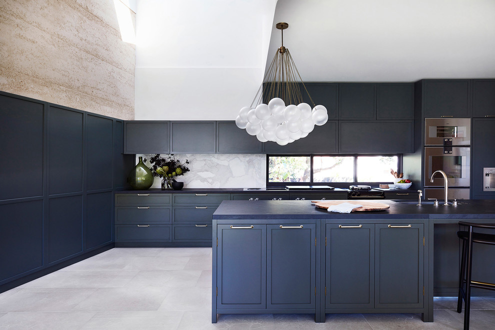 Bild på ett vintage blå blått l-kök, med en undermonterad diskho, skåp i shakerstil, blå skåp, vitt stänkskydd, fönster som stänkskydd, rostfria vitvaror, en köksö och grått golv