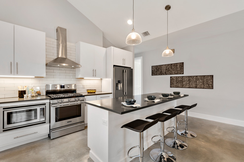 Example of a large minimalist l-shaped concrete floor and gray floor eat-in kitchen design in Austin with flat-panel cabinets, white cabinets, solid surface countertops, white backsplash, subway tile backsplash, stainless steel appliances and an island