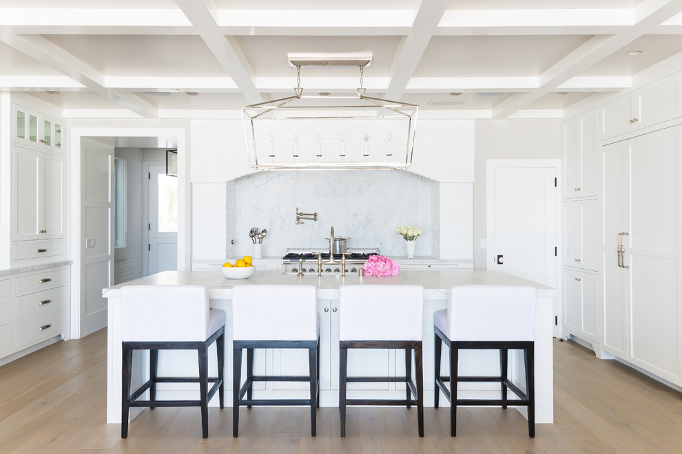 This is an example of a nautical u-shaped kitchen in Orange County with shaker cabinets, white cabinets, white splashback, marble splashback, integrated appliances, medium hardwood flooring, an island and brown floors.