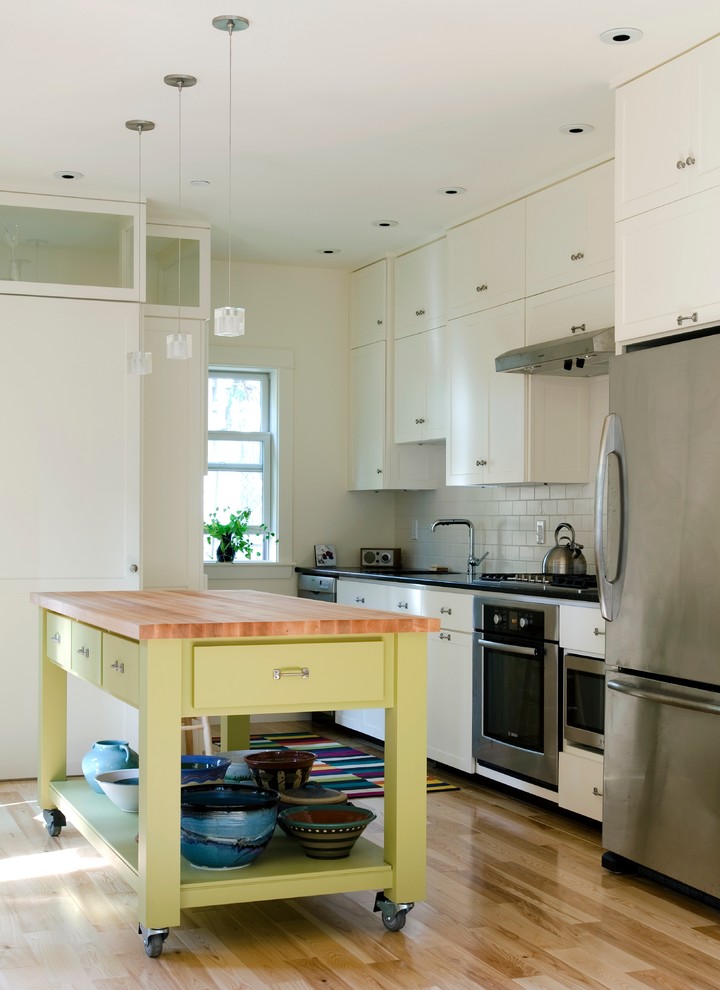 Example of a mid-sized transitional single-wall light wood floor open concept kitchen design in Portland Maine with an integrated sink, recessed-panel cabinets, ceramic backsplash, stainless steel appliances, white cabinets, wood countertops, white backsplash and an island