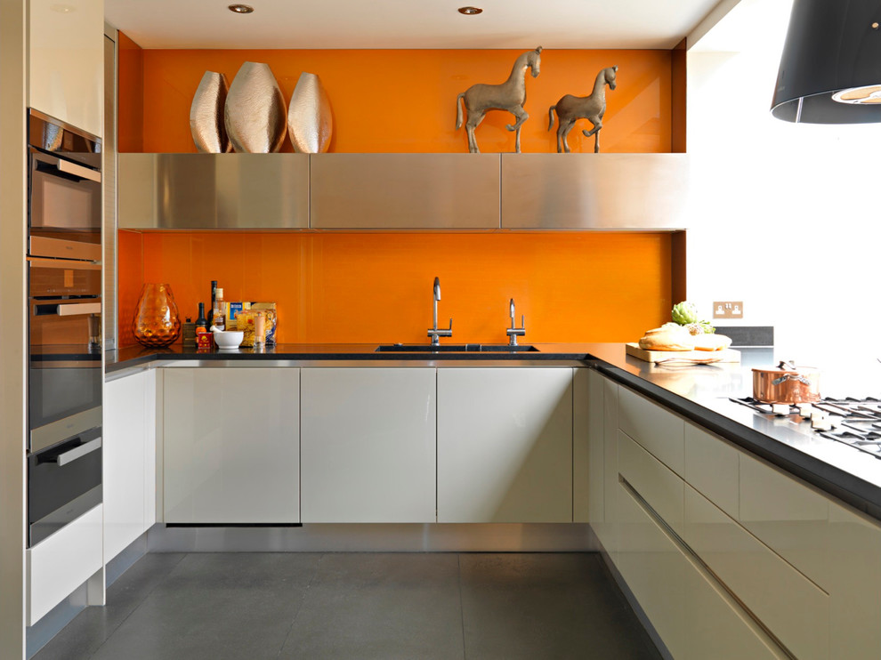 На фото: п-образная кухня в современном стиле с плоскими фасадами, белыми фасадами и оранжевым фартуком с