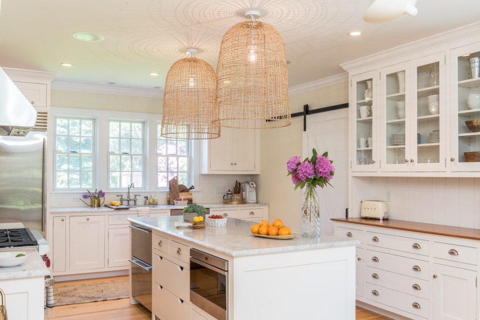 Example of a beach style u-shaped light wood floor kitchen design in New York with a single-bowl sink, glass-front cabinets, white cabinets, wood countertops, white backsplash, stainless steel appliances and an island