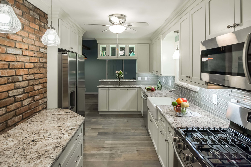 Foto på ett stort kök, med en rustik diskho, luckor med infälld panel, vita skåp, granitbänkskiva, grått stänkskydd, rostfria vitvaror, plywoodgolv och en halv köksö
