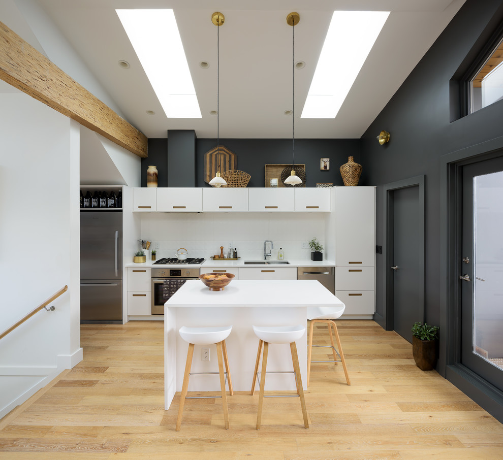 Photo of a scandi single-wall kitchen in Vancouver with a double-bowl sink, flat-panel cabinets, white cabinets, white splashback, stainless steel appliances, light hardwood flooring, an island and beige floors.
