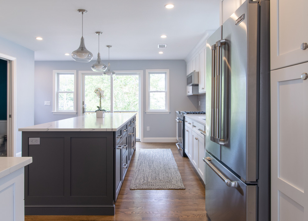 Example of a mid-sized transitional l-shaped medium tone wood floor enclosed kitchen design in New York with an undermount sink, shaker cabinets, white cabinets, quartz countertops, gray backsplash, glass tile backsplash, stainless steel appliances, an island and white countertops
