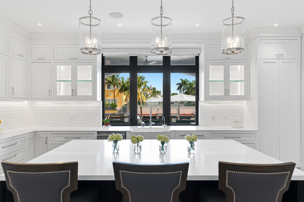 This is an example of an expansive coastal l-shaped kitchen/diner in Miami with white cabinets, engineered stone countertops, white splashback, metro tiled splashback, an island, grey floors, white worktops, a belfast sink, shaker cabinets, stainless steel appliances and porcelain flooring.