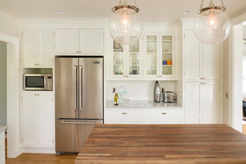 Mid-sized eclectic u-shaped medium tone wood floor eat-in kitchen photo in Portland with a farmhouse sink, shaker cabinets, white cabinets, marble countertops, white backsplash, subway tile backsplash, stainless steel appliances and an island