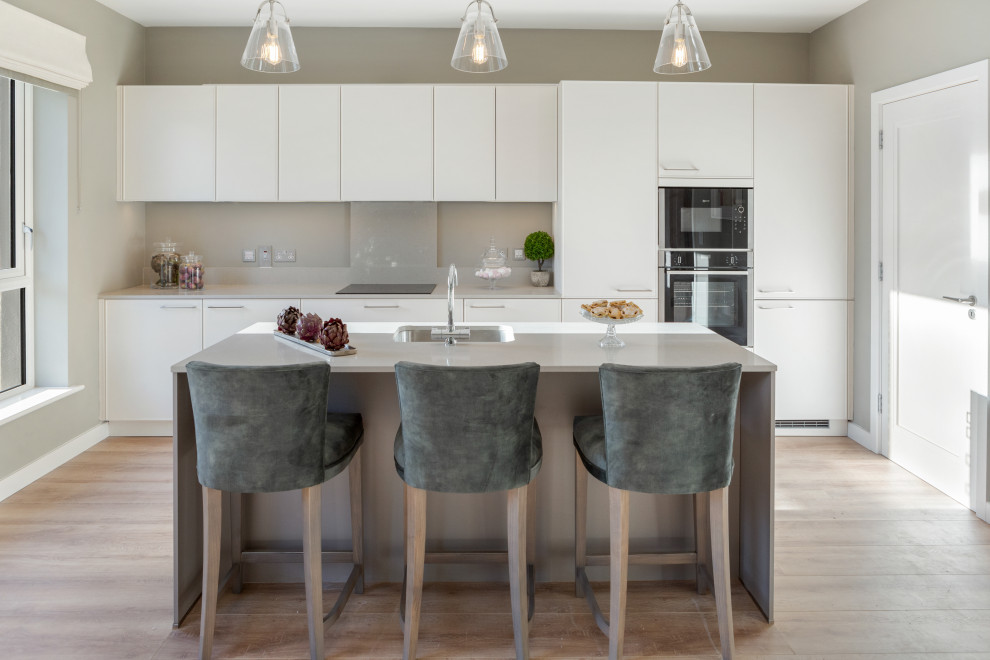 Inspiration for a large contemporary galley kitchen in Dublin with a submerged sink, flat-panel cabinets, white cabinets, grey splashback, integrated appliances, an island, beige floors and grey worktops.