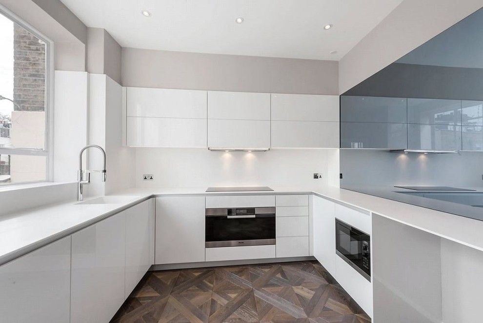 Example of a mid-sized trendy u-shaped medium tone wood floor enclosed kitchen design in London with an undermount sink, flat-panel cabinets, white cabinets, granite countertops, white backsplash, stone tile backsplash, stainless steel appliances and no island