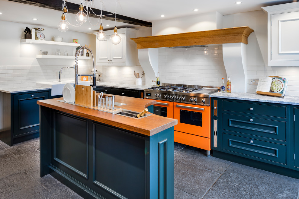 Klassische Küche in L-Form mit Landhausspüle, Kassettenfronten, blauen Schränken, Küchenrückwand in Weiß, Rückwand aus Metrofliesen, bunten Elektrogeräten, Kücheninsel, grauem Boden und Marmor-Arbeitsplatte in Dorset