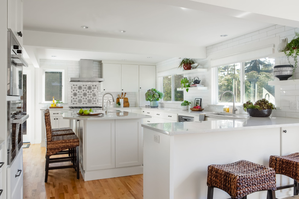 Inspiration for a large eclectic u-shaped kitchen/diner in Seattle with a submerged sink, shaker cabinets, white cabinets, engineered stone countertops, white splashback, porcelain splashback, stainless steel appliances, light hardwood flooring, an island, brown floors and grey worktops.