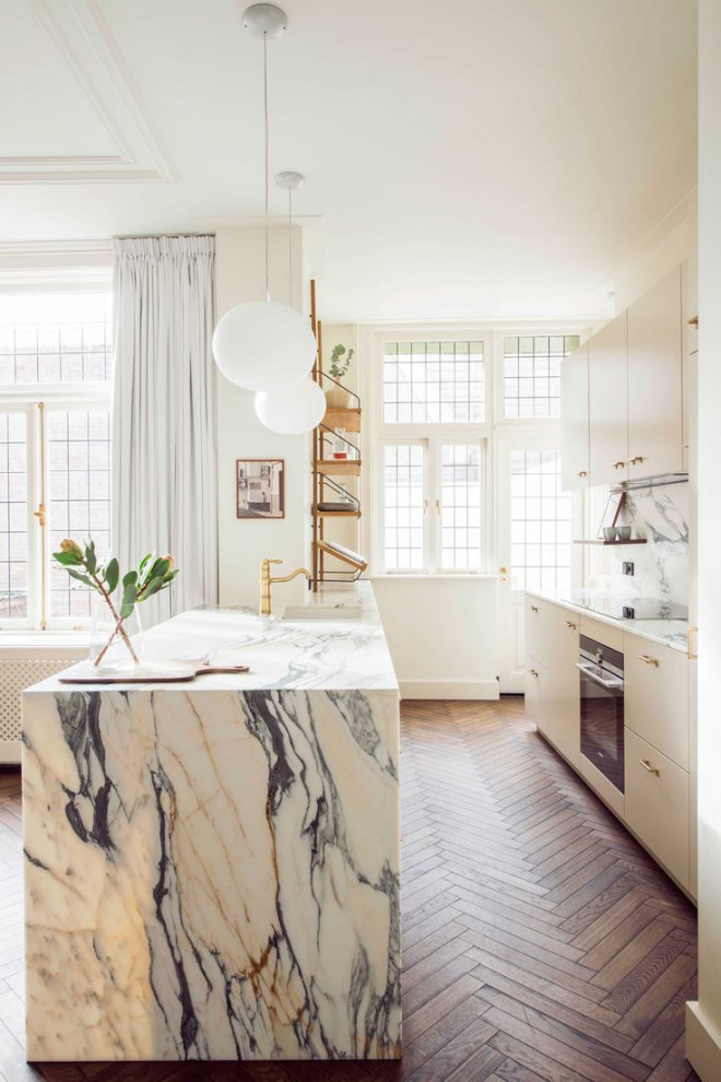 Eklektisk inredning av ett mellanstort vit linjärt vitt kök med öppen planlösning, med en undermonterad diskho, släta luckor, vita skåp, marmorbänkskiva, vitt stänkskydd, stänkskydd i marmor, rostfria vitvaror, mellanmörkt trägolv, en köksö och brunt golv