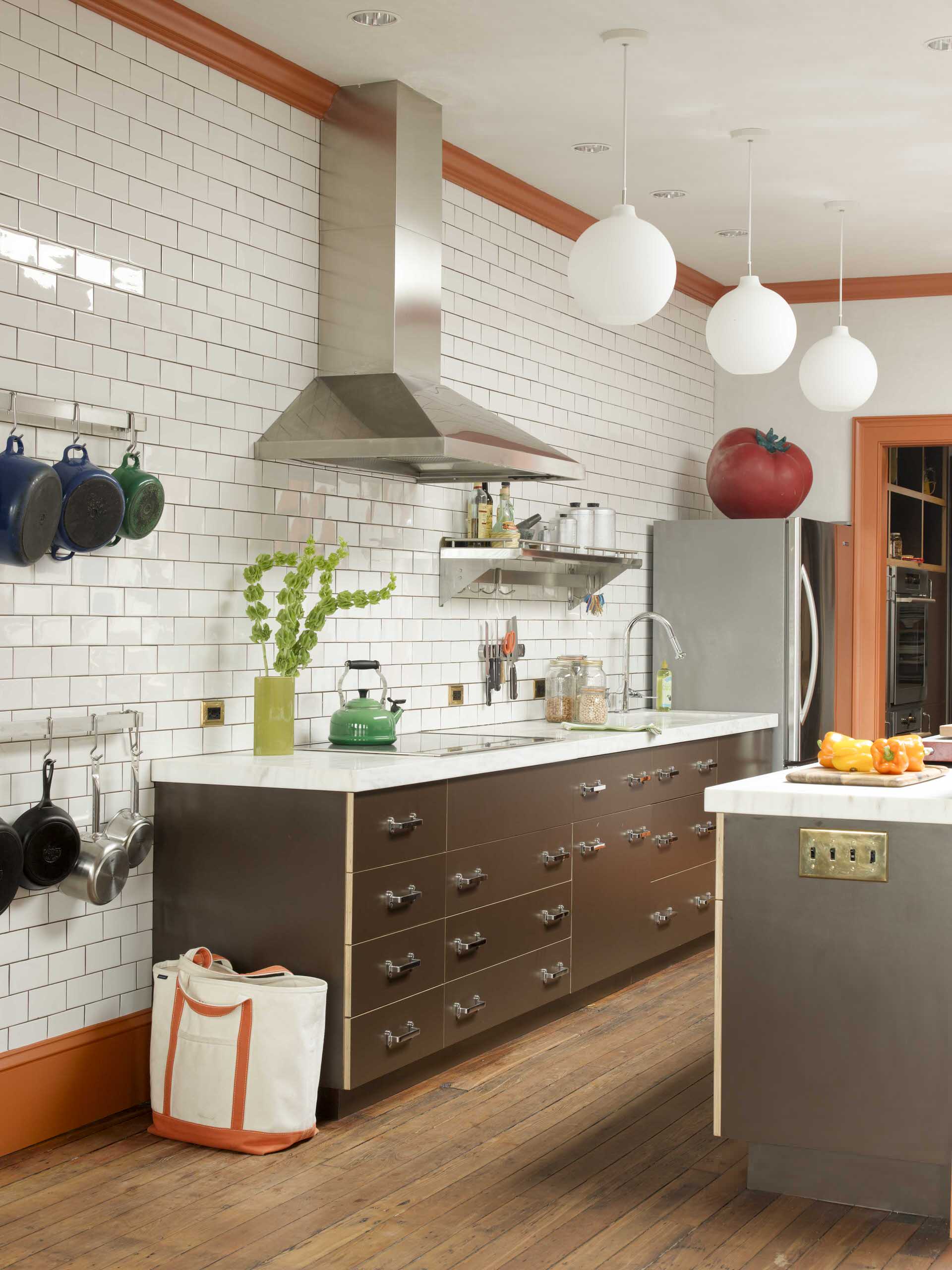 Stacked brown kitchen cabinets flank a black range hood mounted to white  subway backsplash tiles over a stai…