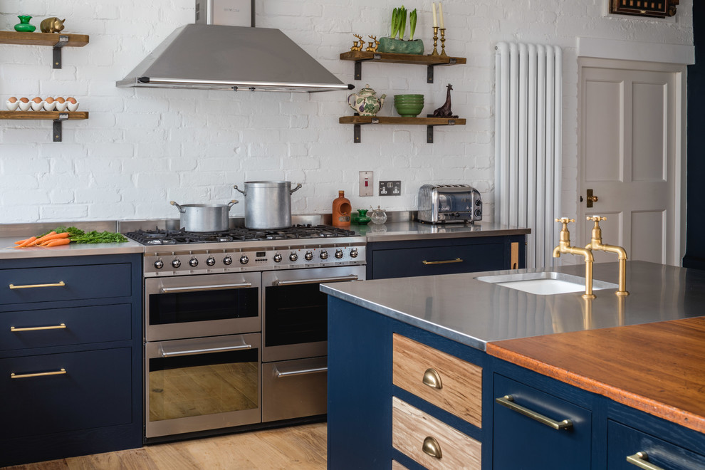 Cette photo montre une cuisine éclectique avec un placard à porte shaker, des portes de placard bleues, un plan de travail en inox, un électroménager en acier inoxydable, un sol en bois brun et îlot.