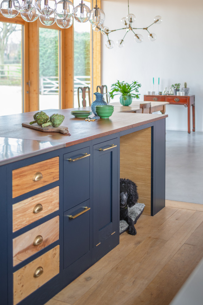 Eklektische Küche mit Schrankfronten im Shaker-Stil, blauen Schränken, Edelstahl-Arbeitsplatte, Küchengeräten aus Edelstahl, braunem Holzboden und Kücheninsel in Oxfordshire
