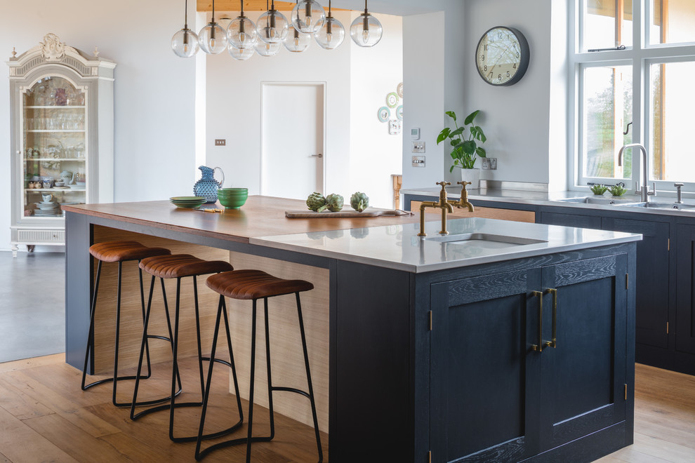 Stilmix Küche mit Schrankfronten im Shaker-Stil, blauen Schränken, Edelstahl-Arbeitsplatte, Küchengeräten aus Edelstahl, braunem Holzboden und Kücheninsel in Oxfordshire