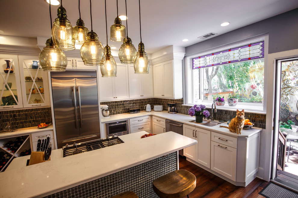 Photo of a medium sized bohemian l-shaped kitchen/diner in Philadelphia with a submerged sink, shaker cabinets, engineered stone countertops, grey splashback, glass tiled splashback, stainless steel appliances, dark hardwood flooring, an island, brown floors and white cabinets.