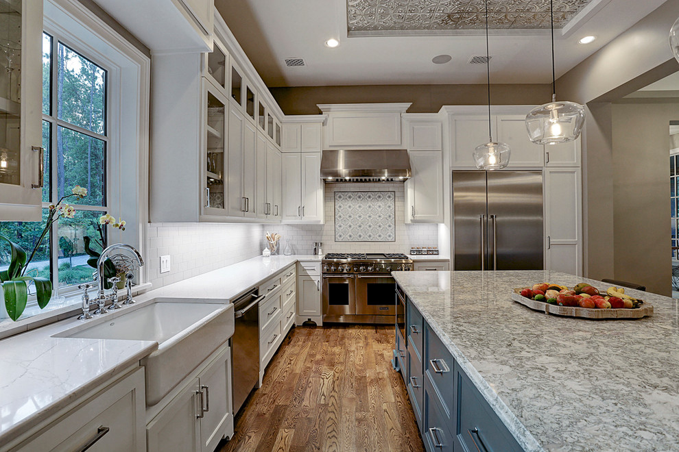 Design ideas for a large farmhouse l-shaped kitchen/diner in Houston with a belfast sink, raised-panel cabinets, white cabinets, granite worktops, white splashback, ceramic splashback, stainless steel appliances, medium hardwood flooring, an island and brown floors.