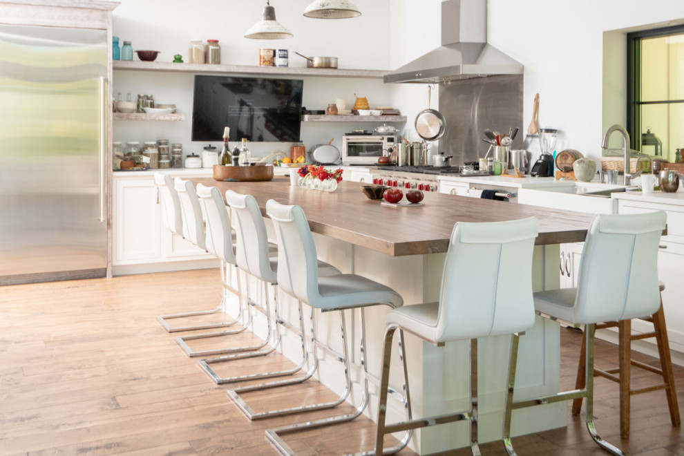 This is an example of an eclectic l-shaped kitchen/diner in Other with a belfast sink, white cabinets, stainless steel appliances, medium hardwood flooring, an island and brown floors.