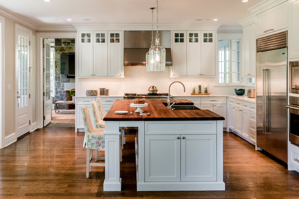 Design ideas for a classic l-shaped kitchen in Chicago with a belfast sink, shaker cabinets, white cabinets, white splashback, stainless steel appliances, dark hardwood flooring, an island, brown floors and wood worktops.