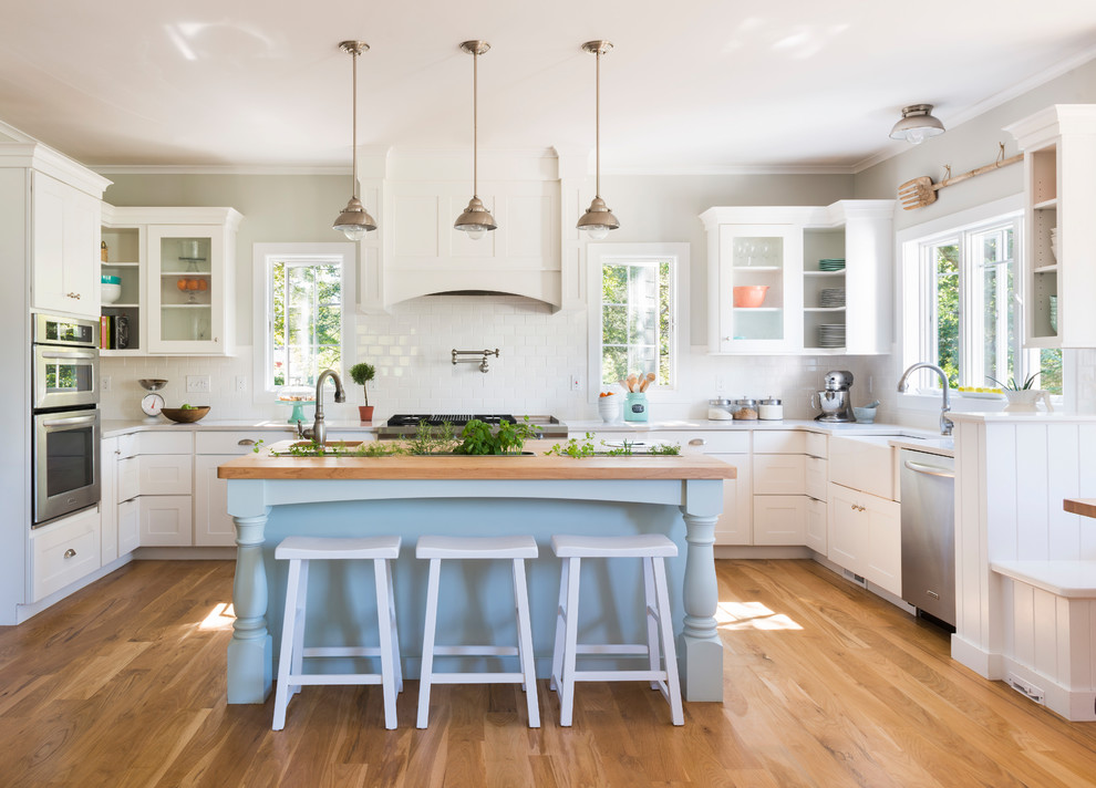 Country u-shaped kitchen in Providence with a belfast sink, shaker cabinets, white cabinets, white splashback, stainless steel appliances, medium hardwood flooring and an island.