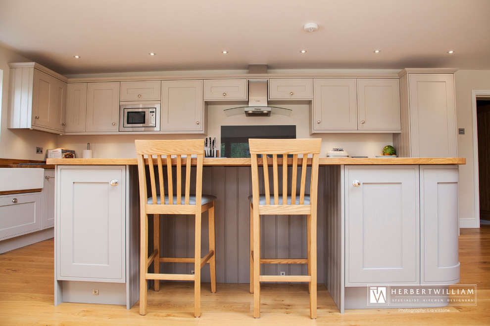 Photo of a country l-shaped open plan kitchen in Hampshire with a belfast sink, shaker cabinets, beige cabinets, wood worktops, integrated appliances, medium hardwood flooring and an island.