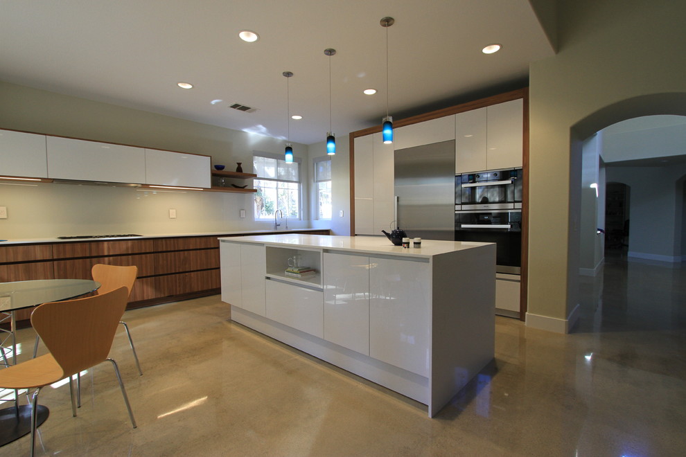 Example of a mid-sized trendy l-shaped concrete floor eat-in kitchen design in Orange County with an undermount sink, flat-panel cabinets, white cabinets, solid surface countertops, white backsplash, glass sheet backsplash, stainless steel appliances and an island