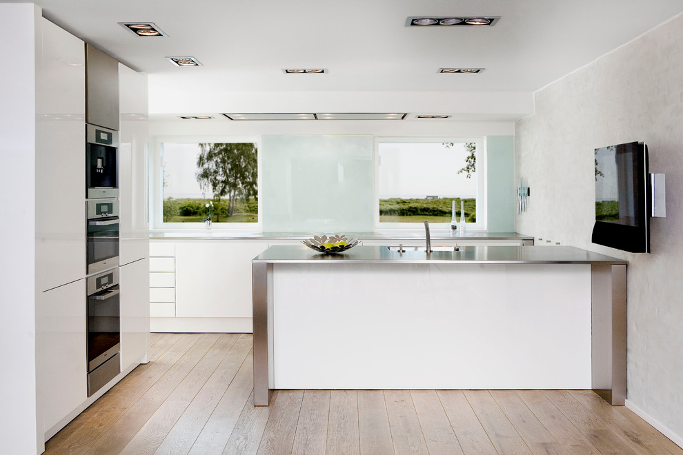 Cette image montre une cuisine parallèle design avec un évier intégré, un placard à porte plane, des portes de placard blanches, un plan de travail en inox, une crédence blanche et une crédence en feuille de verre.